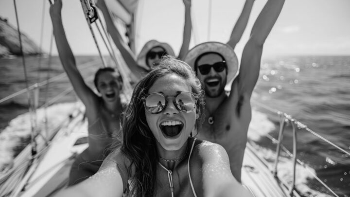 Grupa młodych ludzi na żaglówce, cieszących się żeglowaniem i robiących selfie podczas słonecznego dnia.